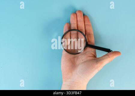 Die Hände des Mannes halten eine Lupe auf dem blauen Tischhintergrund. Der Begriff der sorgfältigen Betrachtung und Suche. Nahaufnahme, POV-Aufnahme. Stockfoto