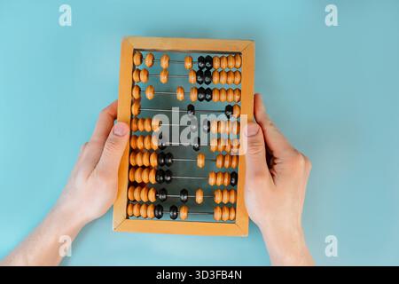 Die Hände des Mannes halten hölzerne alte Abakus auf dem blauen Tischhintergrund. Das Konzept der Buchhaltung und des Geldzählens. Nahaufnahme, POV-Aufnahme. Stockfoto