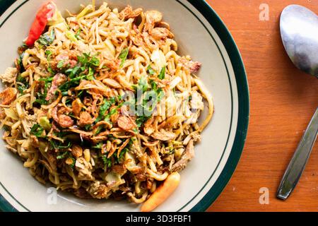 Eine Schüssel mit Instant-Nudeln, garniert mit frischen Kräutern, Chili und frittierten Schalotten, neben einem Löffel. Stockfoto