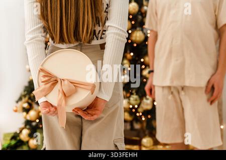 Eine schöne junge Frau hält hinter ihrem Rücken ein Neujahrsgeschenk, ihr Freund steht vor ihr und wartet auf ein Geschenk, ein Zimmer mit einem Weihnachtsbaum Stockfoto