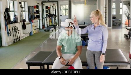 Leitender Trainer in lila Coaching Kunde sitzt auf einer gepolsterten Bank mit VR-Headset im Reha-Fitnessstudio Stockfoto
