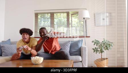 Sie spielen zwei Freunde, die auf einem hellgrauen Sofa im Wohnzimmer sitzen und Controller und Popcorn-Schüssel halten Stockfoto