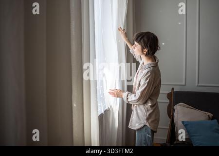 Junge entspannte Frau, die neben dem Fenster im gemütlichen Wohnzimmer stand und morgens weiße Vorhänge öffnete Stockfoto