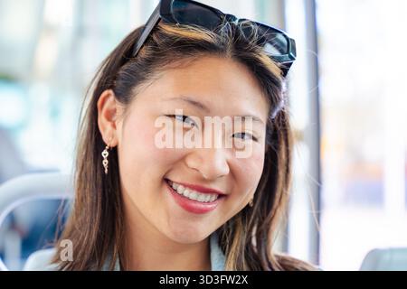 Lächelnde Frau, die in die Kamera blickt und drinnen mit natürlichem Tageslicht sitzt. Stockfoto