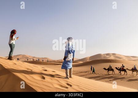 Touristen auf Kamelritten und in traditionellen Tüchern in der Sahara in Merzouga, Marokko, die das Wesen der Berber-Kultur miterleben Stockfoto