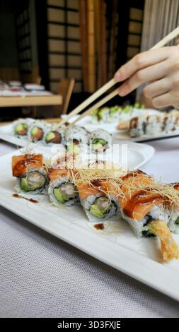 Köstliche Sushi-Brötchen, elegant auf einem weißen Teller angeordnet, garniert mit zarten Garnituren. Eine Hand verwendet meisterhaft Stäbchen, um ein Stück aufzuheben, Showcas Stockfoto