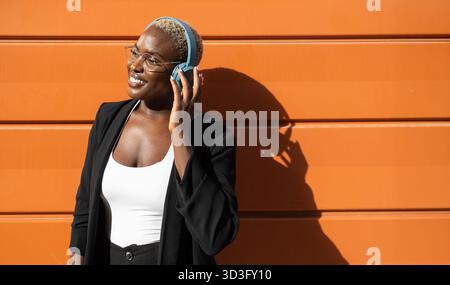 Selbstbewusste Geschäftsfrau und CEO in einem stylischen Blazer lächelt und genießt Musik über Kopfhörer. Sie stand an einer orangen Wand und sah conte aus Stockfoto