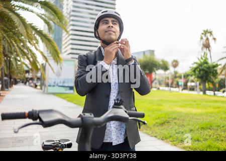 Hispanischer Mann, der einen Helm trägt, während er sich darauf vorbereitet, sein Fahrrad in einem urbanen Park zu fahren. In Geschäftskleidung passt er den Riemen aus Sicherheitsgründen an Stockfoto