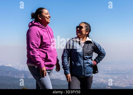 Zwei Frauen lächeln und unterhalten sich während einer Freizeitwanderung an einem sonnigen Tag mit einer atemberaubenden Bergkulisse. Sie scheinen den schönen Sommer zu genießen Stockfoto