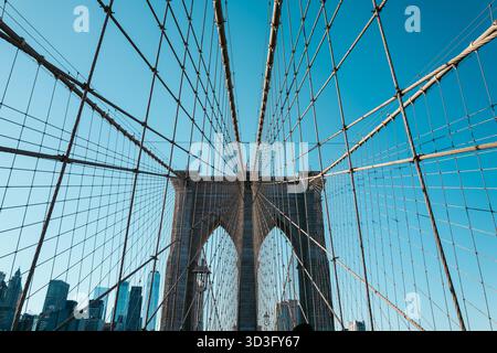 Perspektivische Aussicht auf die berühmte Brooklyn Bridge mit Kabeln, die geometrische Muster vor einem blauen Himmel bilden, und New York City Wolkenkratzer, die im sichtbar sind Stockfoto