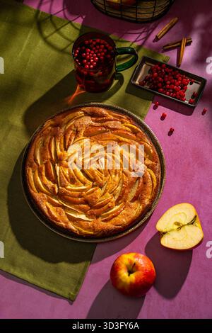Ein wunderschöner Apfelkuchen wird von oben auf einem lebendigen Tisch präsentiert, begleitet von frischen Äpfeln, Preiselbeeren und Zimtstangen, die eine gemütliche Atmosphäre schaffen Stockfoto