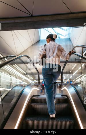 Eine Frau in lässiger Kleidung steigt in einer modernen Kopenhagener U-Bahn-Station eine Rolltreppe hinauf und verkörpert Reisen und urbane Routine. Die schlanke Architektur verbessert Stockfoto