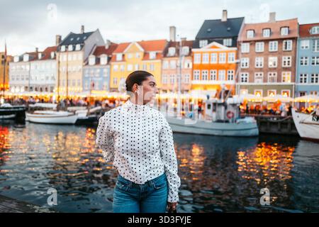 Eine stilvolle Frau steht am Nyhavn-Kanal in Kopenhagen und genießt die lebhafte abendliche Atmosphäre. Farbenfrohe Gebäude und Reflexionen auf dem Wasser Stockfoto