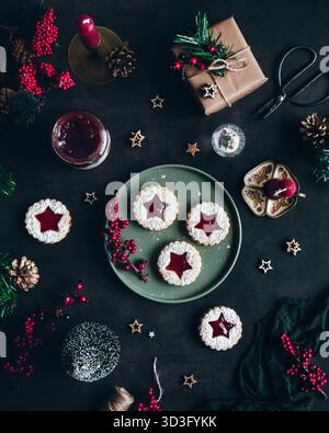 Blick von oben auf sternförmige Marmeladenkekse auf einer grünen Tafel mit festlicher Dekoration. Enthält rote Kerzen, verpacktes Geschenk, Beeren und Grün auf einem dunklen Sur Stockfoto