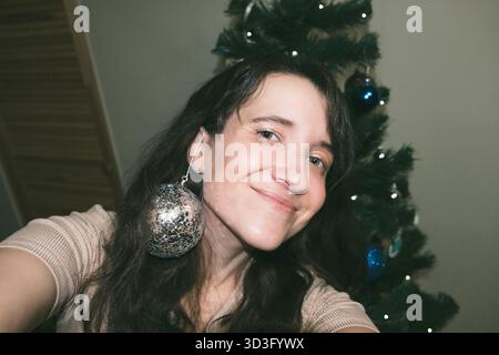 Eine fröhliche Frau posiert für ein Selfie spielerisch vor einem Weihnachtsbaum und trägt ein funkelndes Ornament als Ohrring. Festliche Lichter und Dekorationen e Stockfoto
