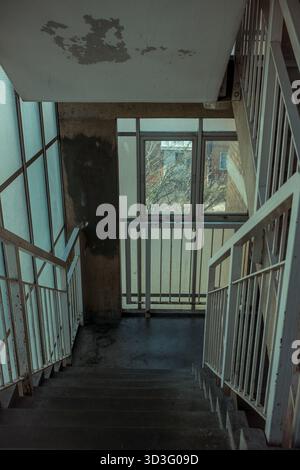 Eine schwach beleuchtete Treppe in einem verlassenen Gebäude. Die abblätternden Wände und die rostigen Geländer deuten auf jahrelange Vernachlässigung hin, während das Fenster einen Blick auf das Fenster bietet Stockfoto