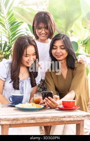 Gruppe asiatischer Teenager, die einen freudigen Moment gemeinsam in einem indonesischen Café im Freien genießen und Fotos auf einem Smartphone teilen, während sie von üppigem Gier umgeben sind Stockfoto