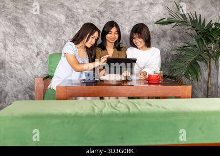 Eine Gruppe asiatischer Teenager sitzt in einem indonesischen Café, genießt Kaffee und benutzt gemeinsam ein Tablet. Die gemütliche Atmosphäre wird durch ein lebhaftes Grün verstärkt Stockfoto