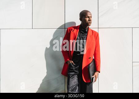 Ein junger LGBTQ-Mann strahlt Vertrauen in eine leuchtend rote Jacke aus, die sich an eine moderne, helle Wand lehnt. Er schaut nachdenklich in die Ferne. Stockfoto
