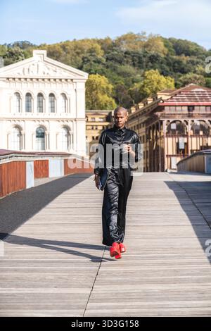 Ein junger LGBTQ-Mann geht selbstbewusst auf einem Holzdeck, trägt stilvolle schwarze Kleidung und auffällige rote Absätze. Der Hintergrund ist ein klassisches Bauwerk Stockfoto