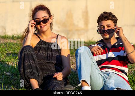 Ein Paar genießt einen sonnigen Tag im Freien, sitzt auf grasbewachsenem Boden und trägt eine stilvolle Sonnenbrille. Ihr lässiger Stil und ihr entspanntes Auftreten betonen ein Auto Stockfoto