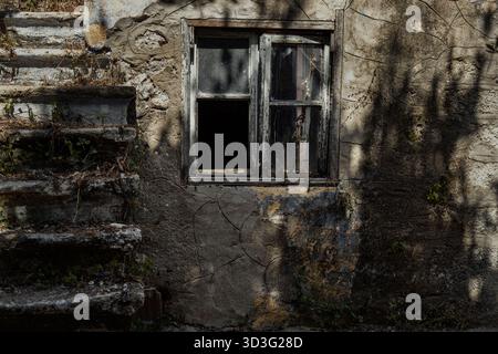 Eine alte Steinmauer mit einem verwitterten Holzfenster und einer alten Treppe. Schatten erzeugen eine geheimnisvolle Atmosphäre, die die Textur und die Geschichte hervorhebt Stockfoto