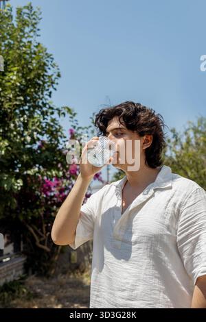 Ein junger Mann mit geschlossenen Augen trinkt an einem sonnigen Tag Wasser aus einem Glas. Er steht draußen und bleibt unter dem klaren blauen Himmel, umgeben von Gr Stockfoto