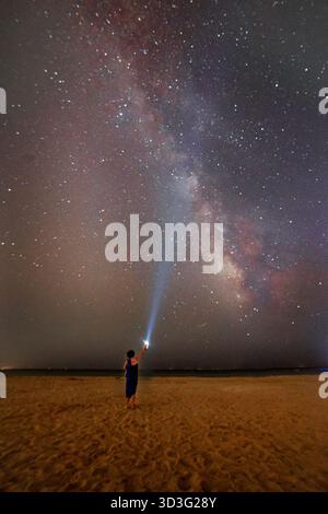 Ein Mann steht an einem Sandstrand unter der Weite der Milchstraße. Er zeigt eine Taschenlampe in Richtung Himmel und schafft eine auffällige Verbindung zwischen Stockfoto