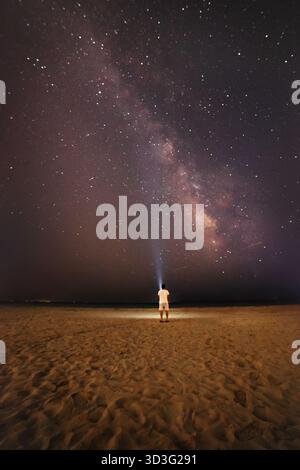 Ein einsamer Mann steht an einem Sandstrand und blickt nach oben auf die weitläufige Milchstraße. Der Nachthimmel ist voller Sterne, die eine faszinierende und ruhige Atmosphäre schaffen Stockfoto