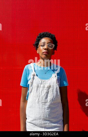 Eine Frau mit blauem Hemd und weißem Overall steht selbstbewusst vor einer leuchtend roten Wand. Der helle Hintergrund verbessert den Kontrast, übertragen Stockfoto