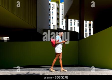 Eine Frau mit rotem Rucksack läuft, während sie ihr Telefon benutzt, umgeben von moderner Architektur in einer Stadt. Das helle Sonnenlicht wirft scharfe Schatten, hochglänzend Stockfoto