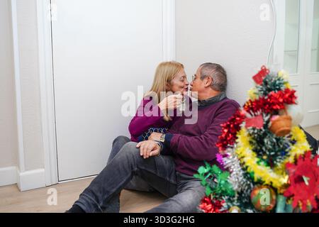 Ein reifes Paar teilt einen intimen Moment bei einem wunderschön dekorierten Weihnachtsbaum, der die Essenz festlicher Wärme, Liebe und Urlaubstradition einfängt Stockfoto