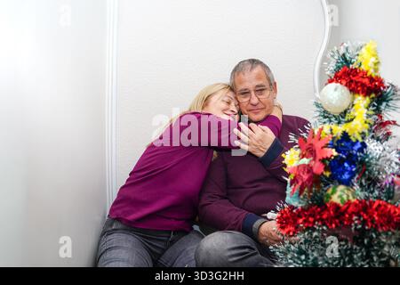Ein reifes Paar in festlichen Pullover sitzt liebevoll neben einem wunderschön geschmückten Weihnachtsbaum und verkörpert die Wärme und Freude der Urlaubstraditionen Stockfoto