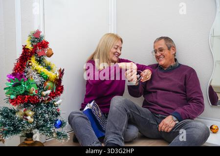 Ein freudiges, reifes Paar teilt einen herzzerreißenden Moment, sitzt an einem wunderschön geschmückten Weihnachtsbaum, tauscht Geschenke aus und genießt den festlichen Urlaub Stockfoto