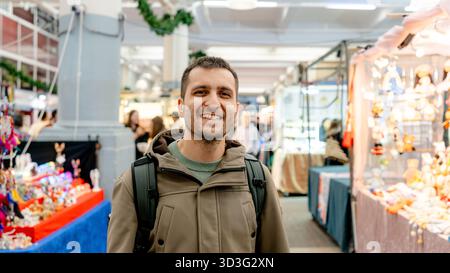 Ein glücklicher Mann mit einem Rucksack, der warm in die Kamera lächelt, während er die pulsierenden Verkaufsstände am Covent Garden Market in London erkundet und den lebhaften geldautomaten genießt Stockfoto