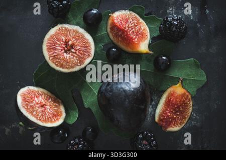 Frische dunkle Feigen, Heidelbeeren und Brombeeren, wunderschön auf grünen Blättern mit dunklem Hintergrund angeordnet Stockfoto