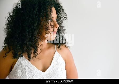 Ein ruhiges Porträt einer Frau mit voluminösen lockigen Haaren, die nachdenklich zur Seite schaut, gekleidet in einem eleganten weißen Spitzenkleid. Ein dezentes weißes Backgr Stockfoto