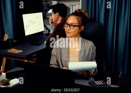 Geschäftsfrau, die nach draußen blickt und die Datenanalyse durchdenkt, überlastete die Projektarbeit, während sie eine Brille mit einem Kollegen hielt, der hinten bei arbeitete Stockfoto