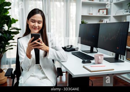 Lächelnde junge asiatische Geschäftsfrau mit glücklichem Gesicht, die auf Smartphone in profitablen Börsenkurven in modernen Büros schaut. Konzept von Stockfoto