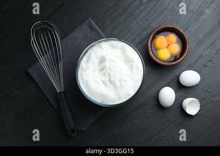 Eiweiß in Schüssel, Schneebesen und Zutaten auf schwarzem Tisch, flach gelegt Stockfoto
