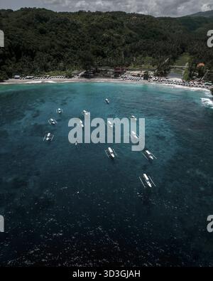 Aus der Vogelperspektive sehen Sie Boote, die auf kristallklarem türkisfarbenem Wasser schwimmen und auf die Sandküste unter einem üppigen grünen Hügel treffen, Bali, Indonesien. Stockfoto