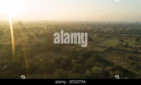 Aus der Vogelperspektive schweben Heißluftballons ruhig über antike Tempel und üppige Landschaften, die in das sanfte, goldene Licht der Morgenröte getaucht sind, Old Bagan, Mandalay Stockfoto