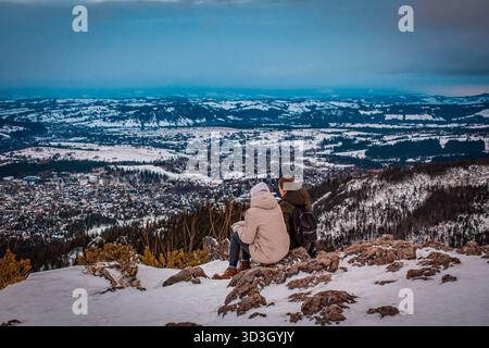 Zwei Mädchen ruhen sich auf einem Berggipfel aus, umgeben von einer atemberaubenden Winterlandschaft mit Blick auf den Wald und die schneebedeckten Gipfel. Stockfoto