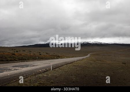 Einsame Straße, die sich unter stürmischem Himmel durch den hohen páramo schlängelt und zu den schneebedeckten Hängen des Monte Chimborazo in Ecuador führt. Stockfoto