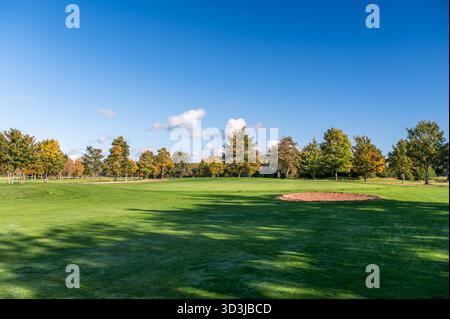 Weite Aussicht auf ein Golfgrün mit Flagge, umgeben von Bäumen, die Herbstfarben unter einem klaren blauen Himmel zeigen. Die Szene zeigt hellgrünes Gras und ein ruhiges Ou Stockfoto