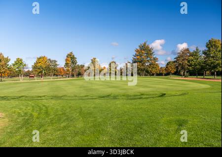 Weite Aussicht auf ein Golfgrün mit Flagge, umgeben von Bäumen, die Herbstfarben unter einem klaren blauen Himmel zeigen. Die Szene zeigt hellgrünes Gras und ein ruhiges Ou Stockfoto