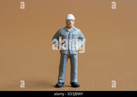 Miniaturbauarbeiter. Ein Mann in einer blauen Arbeitsuniform, komplett mit einem Schutzhelm. Er steht selbstbewusst mit den Händen auf den Hüften, Proje Stockfoto