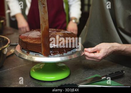 Essen, Nahaufnahme einer Person, die während der Dessertzubereitung auf einem rotierenden Kuchenständer reichhaltige Schokoladenganache auf einen Kuchen glättet Stockfoto