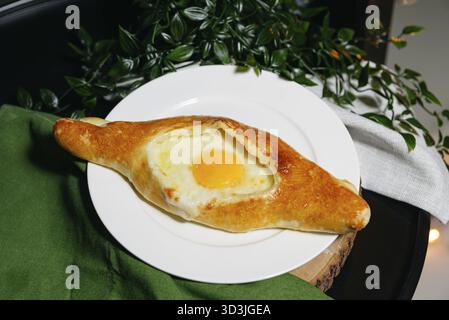 Essen, frisch gebackener adjarischer Khachapuri gefüllt mit geschmolzenem Käse und sonnigem Eigelb, serviert auf einem weißen Teller mit Kräutern Stockfoto