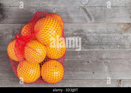 Hellgelbe Zitrusfrüchte in einem roten Netzbeutel auf rustikalen Holzbrettern. Stockfoto